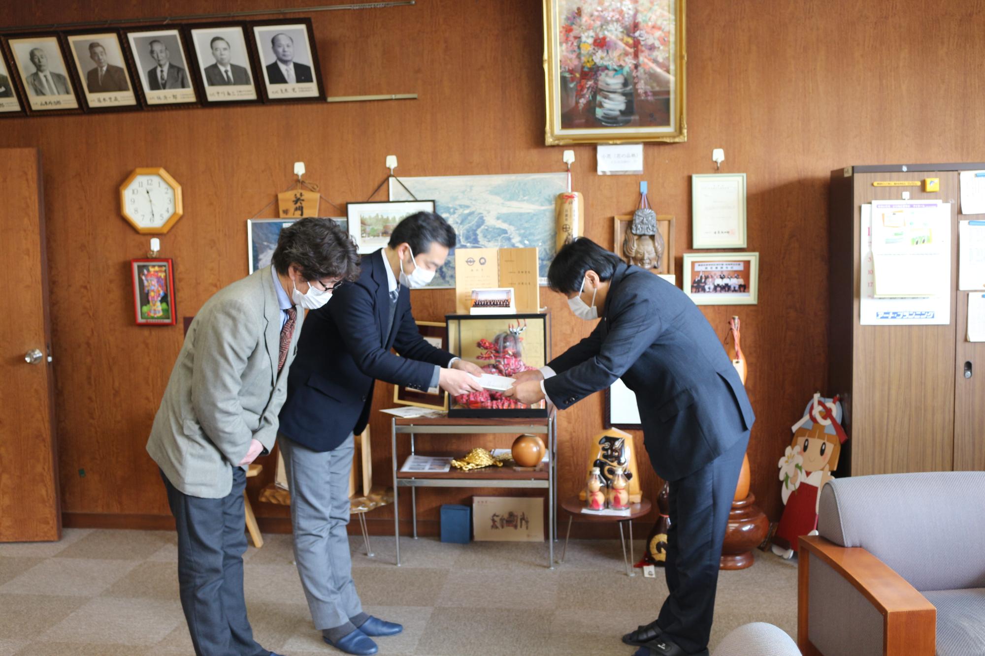 スーツ姿の二人組の男性がもう一人のスーツ姿の男性にお辞儀しながら紙を渡し、それをお辞儀しながら写真